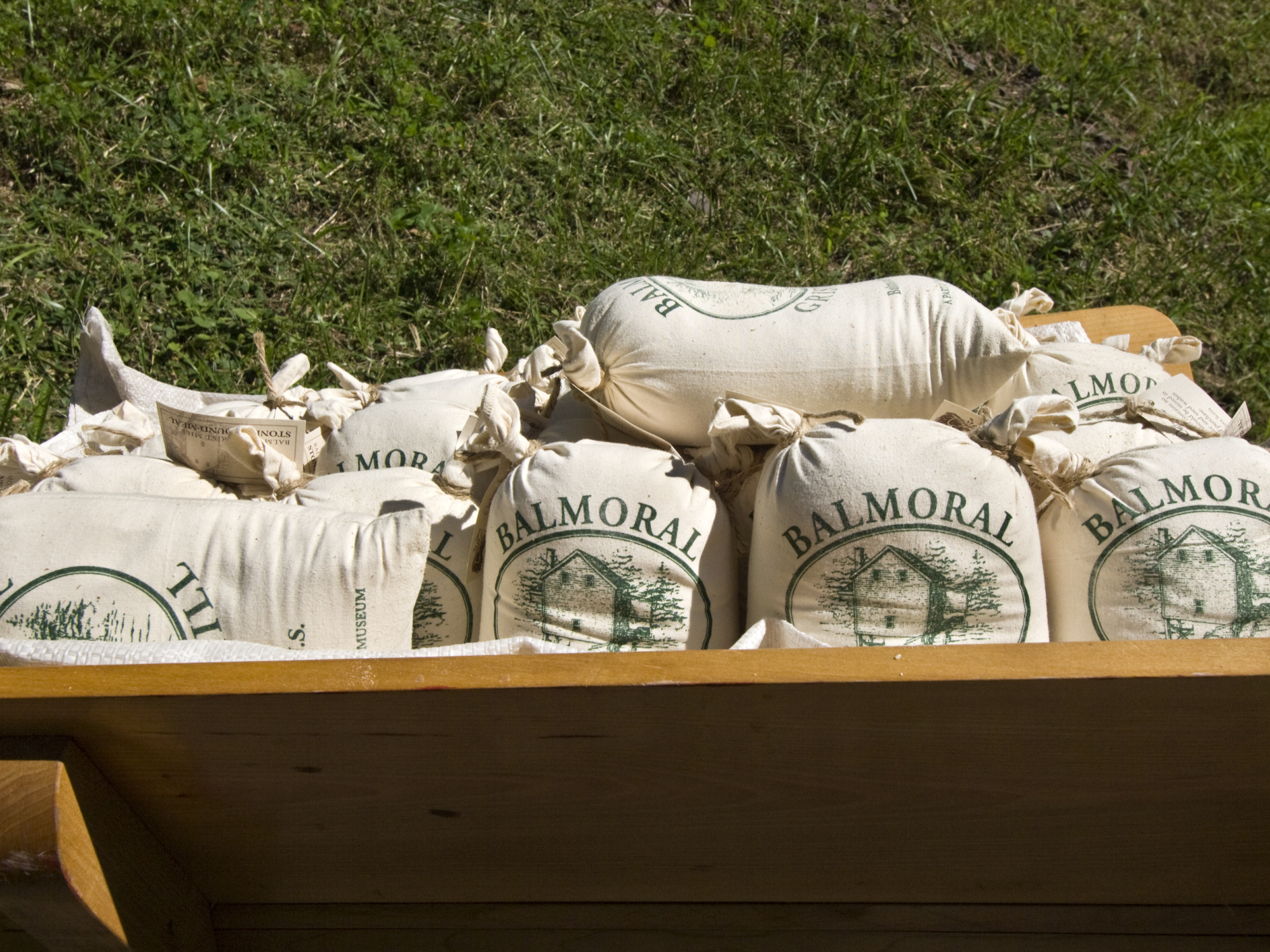 Several white cloth sacks filled with flour sit on a wooden wheelbarrow with green grass visible in the background. The sacks are labeled 'Balmoral' and feature an illustration of a building